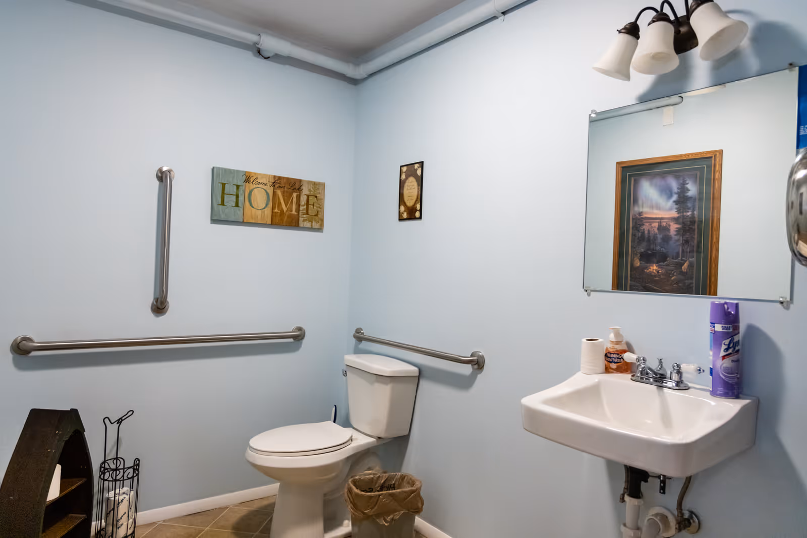 A clean bathroom with light blue walls featuring a white toilet, a white sink with a mirror above it, and stainless steel grab bars on the walls. There is a small trash can next to the toilet, a decorative sign on the wall that says 'Welcome Home', and various toiletries on the sink including a roll of toilet paper, hand soap, and a can of Lysol disinfectant spray. A framed picture is reflected in the mirror.