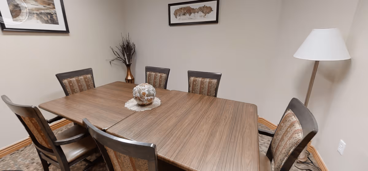 A small dining area with a rectangular wooden table surrounded by six chairs with patterned upholstery. On the table is a decorative spherical object placed on a lace doily. The room has beige walls with two framed pictures hanging, a tall floor lamp in the corner, and a vase with dried branches on a small stand.