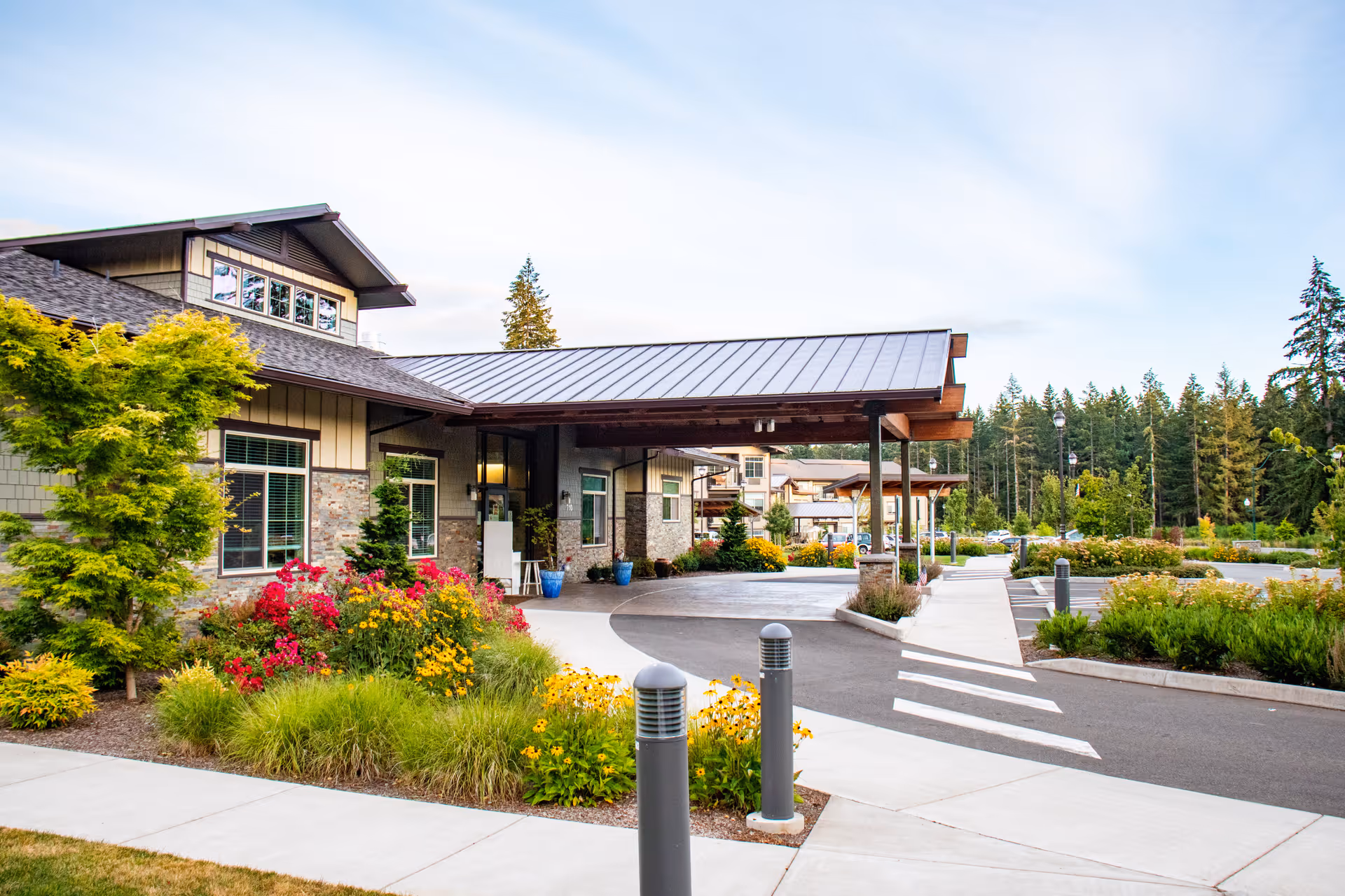 Exterior view of Fieldstone Memory Care Olympia facility showing the entrance with a covered drop-off area, landscaped garden with colorful flowers and shrubs, a paved driveway, and a parking area surrounded by trees under a partly cloudy sky.