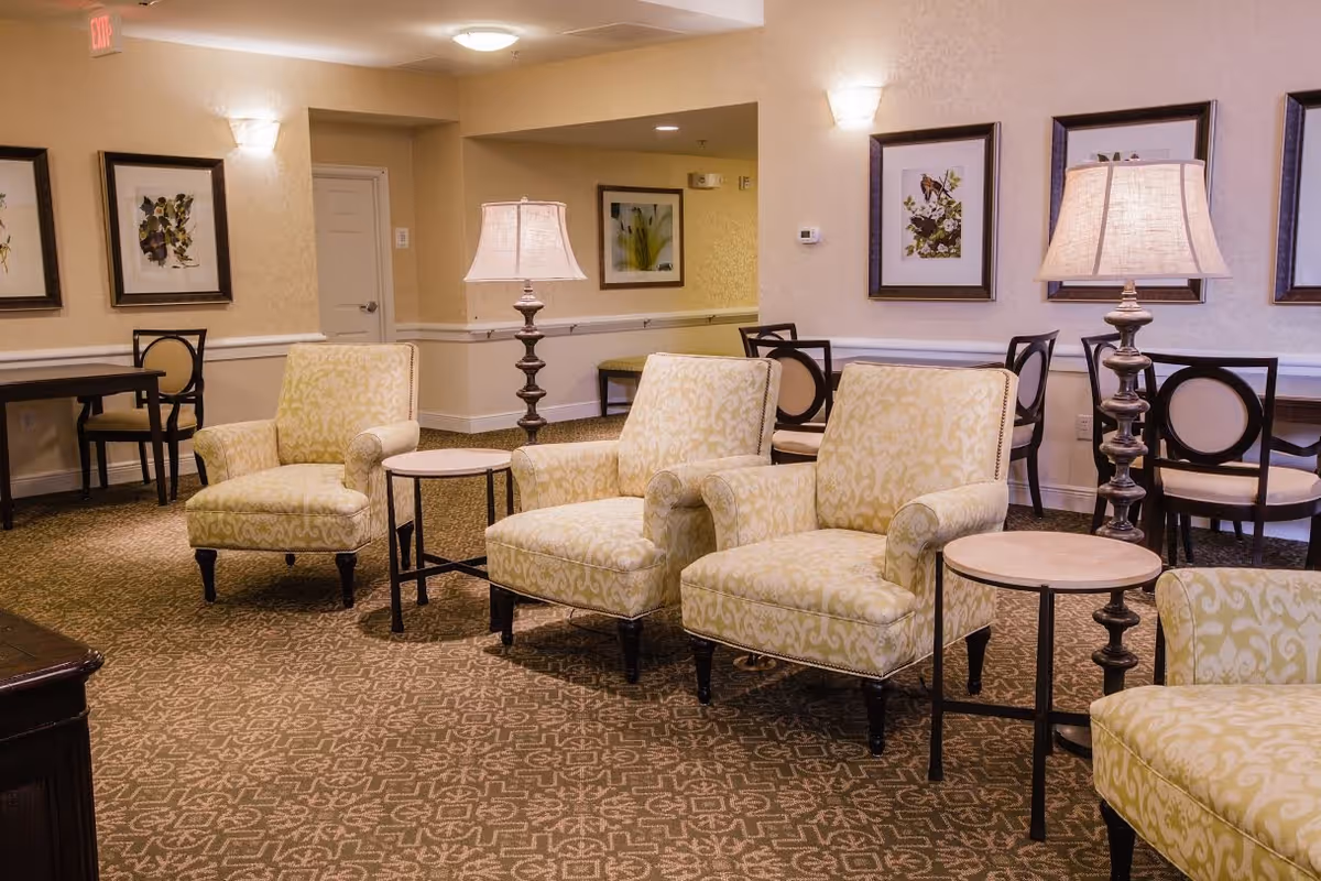 A cozy senior living common area with patterned beige armchairs arranged around small round tables, decorative table lamps, framed botanical artwork on the walls, and additional seating with dark wood chairs and tables in the background.