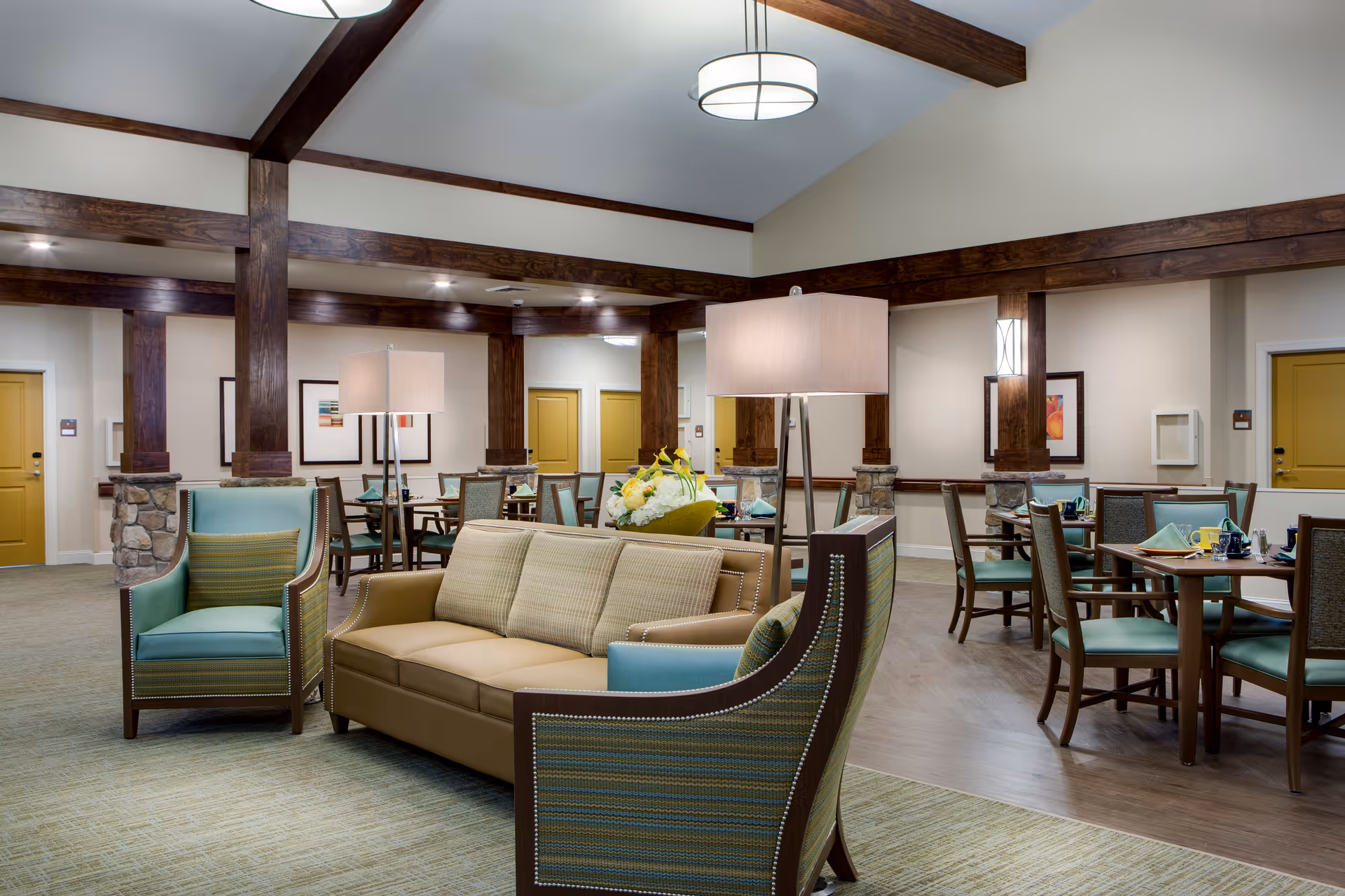 A spacious and well-lit common area in a senior living facility featuring a beige sofa and two green armchairs with patterned cushions. The room has wooden beams and columns with stone bases, multiple round dining tables with chairs, and table settings including napkins and cups. The walls are decorated with framed artwork, and there are several yellow doors visible in the background.