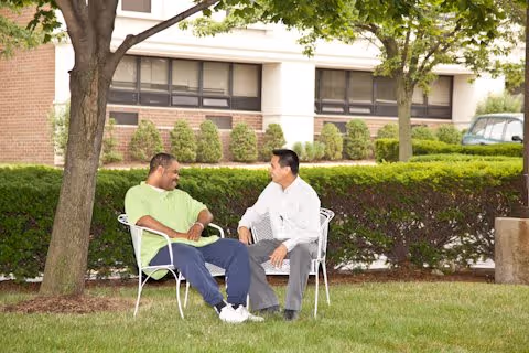 Two men sitting on white metal chairs on a grassy area outside, engaged in conversation. They are near trees and bushes with a building visible in the background.