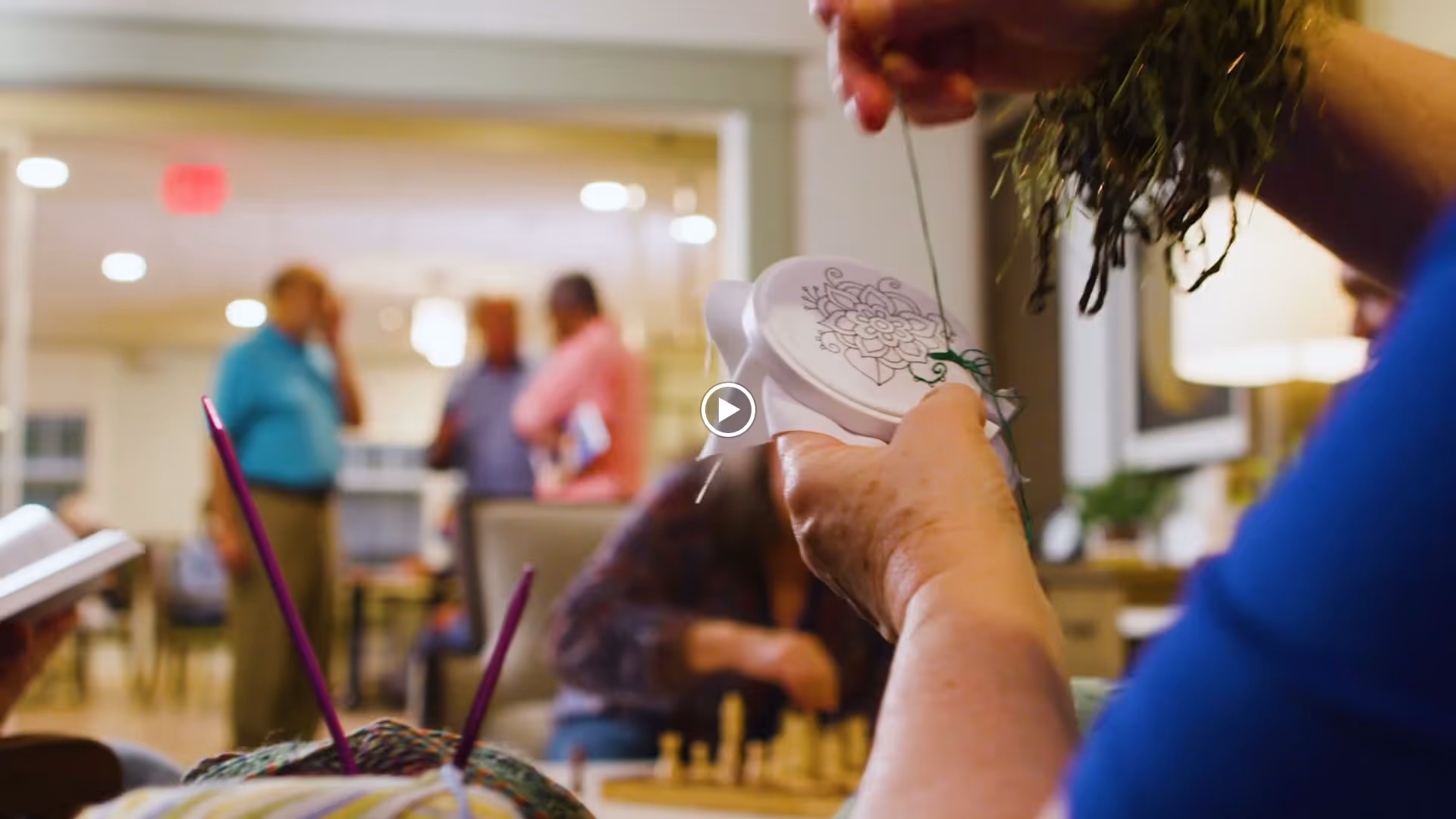 Close-up of a person holding an embroidery hoop and needle, working on a floral design. In the background, several people are engaged in conversation and activities in a well-lit common area of a senior living facility.