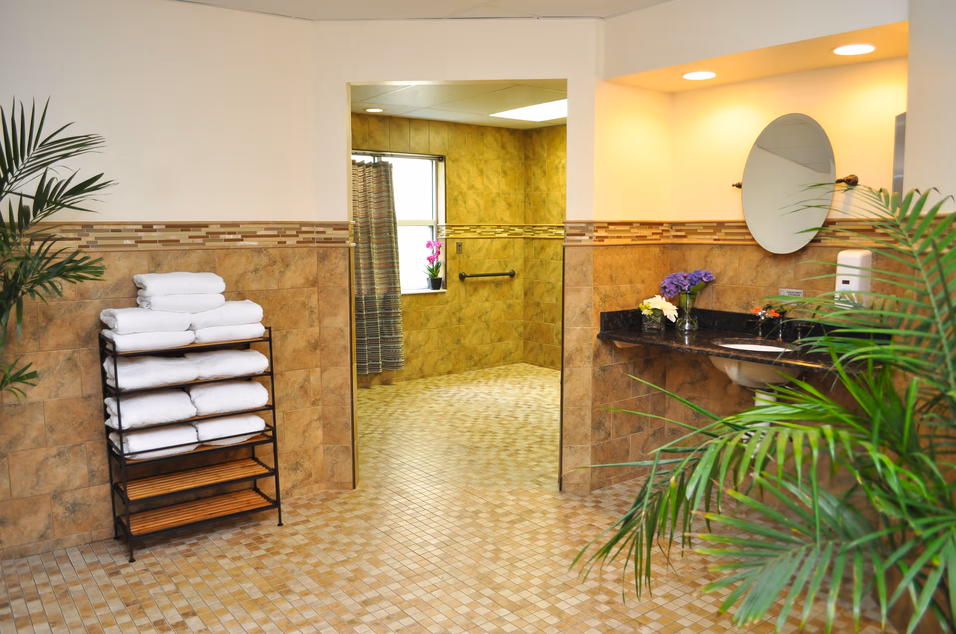A clean and spacious bathroom area with tiled floors and walls. On the left, there is a rack with neatly folded white towels. In the center, an open doorway leads to a shower area with a window and a shower curtain. On the right, there is a countertop with a sink, a round mirror above it, a soap dispenser, and decorative flowers. Green plants are placed on both sides of the room, adding a touch of nature.