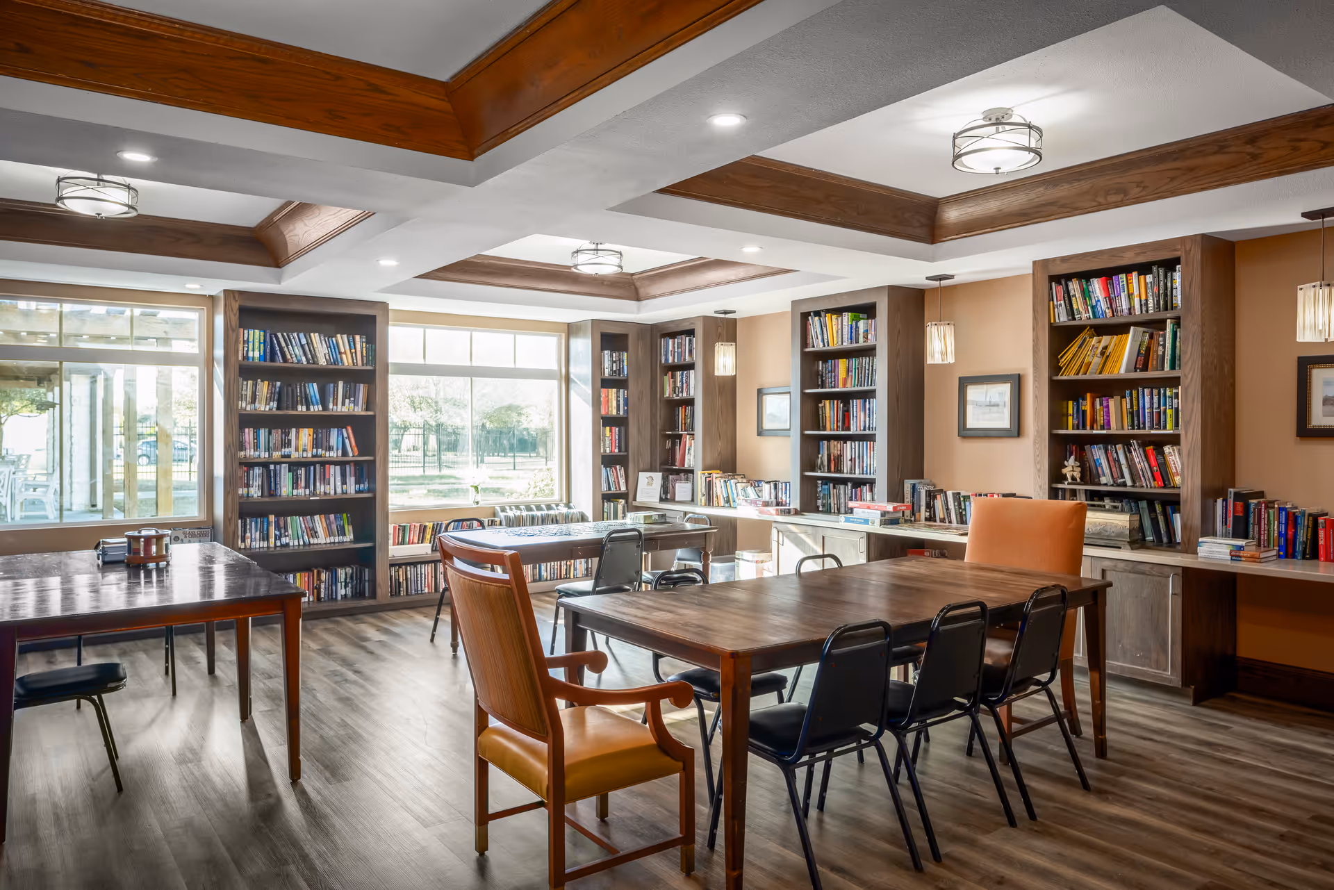A bright and spacious library or reading room with large windows letting in natural light. The room features wooden bookshelves filled with books, wooden tables with chairs around them, and a ceiling with wooden beams and modern light fixtures. The floor is wood, and the walls are painted a warm beige color.