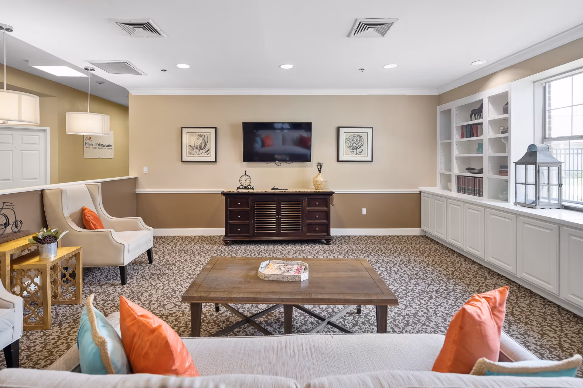 A cozy living room area in a senior living facility with a beige and brown color scheme. The room features a flat-screen TV mounted on the wall above a dark wooden cabinet, two framed botanical prints on either side of the TV, a large window with white built-in shelves and cabinets beneath it, and comfortable seating including a sofa with colorful pillows and an armchair with an orange pillow. There is a wooden coffee table in the center with a decorative tray on top, and a small side table with a plant and decorative items.