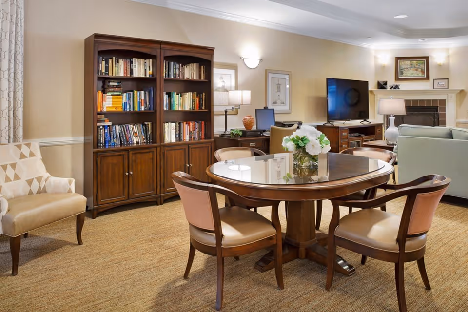 A cozy living area with a round wooden table surrounded by four chairs, a wooden bookshelf filled with books, a beige armchair, a desk with a computer, a flat-screen TV on a wooden stand, and a fireplace with a painting above it. The room has warm lighting and neutral-colored walls and carpet.