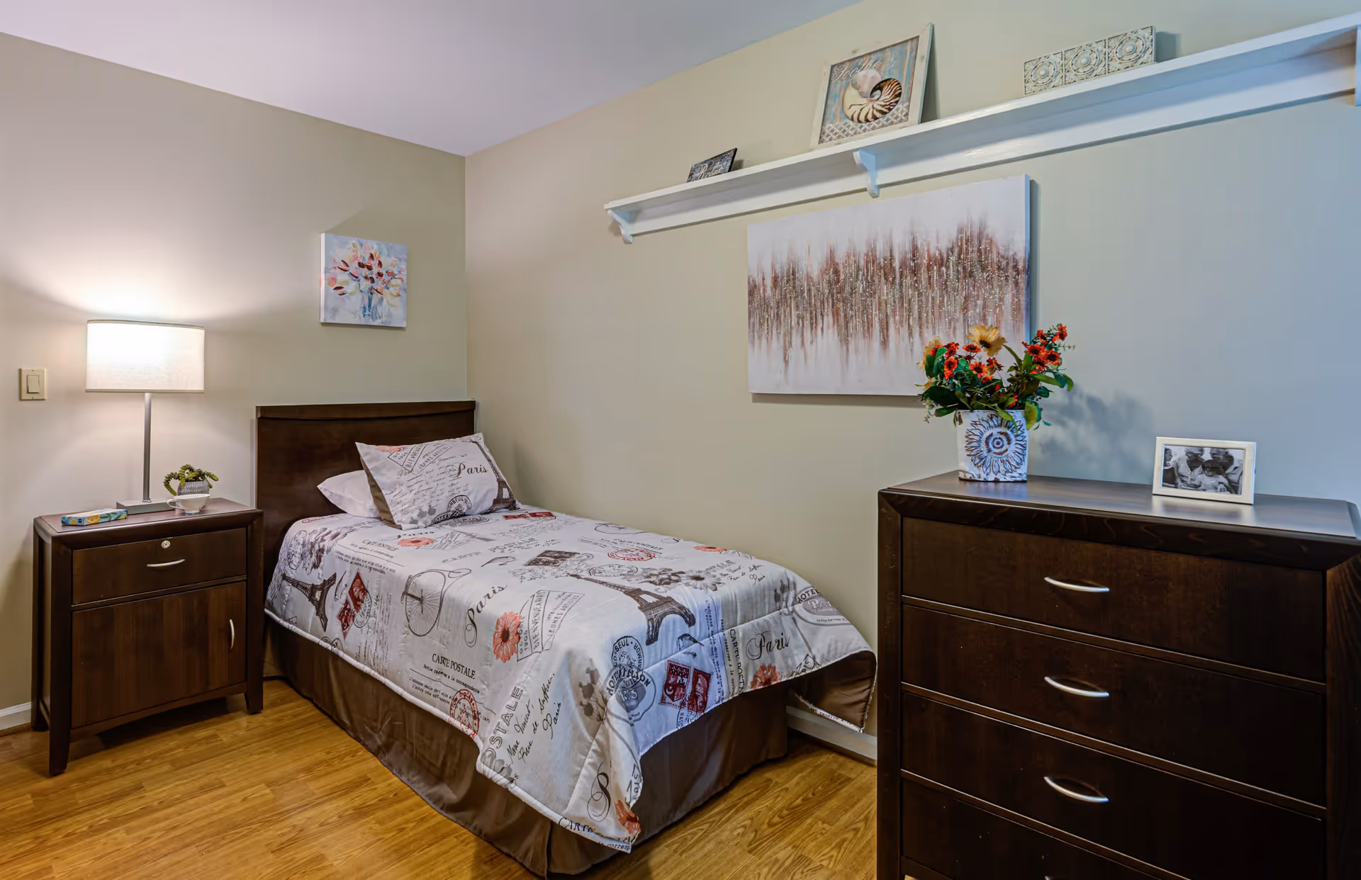 A small bedroom with a single bed covered in a Paris-themed bedspread and pillow. Next to the bed is a wooden nightstand with a lamp, a small plant, and a book. Across from the bed is a dark wooden dresser with three drawers, decorated with a vase of flowers and a framed photo. The walls are light beige with two pieces of artwork hanging above the bed and dresser. The floor is wooden.