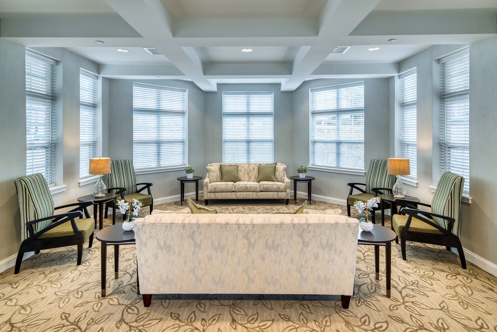 Bright communal living room with sofas, striped armchairs, side tables, lamps, and large windows.