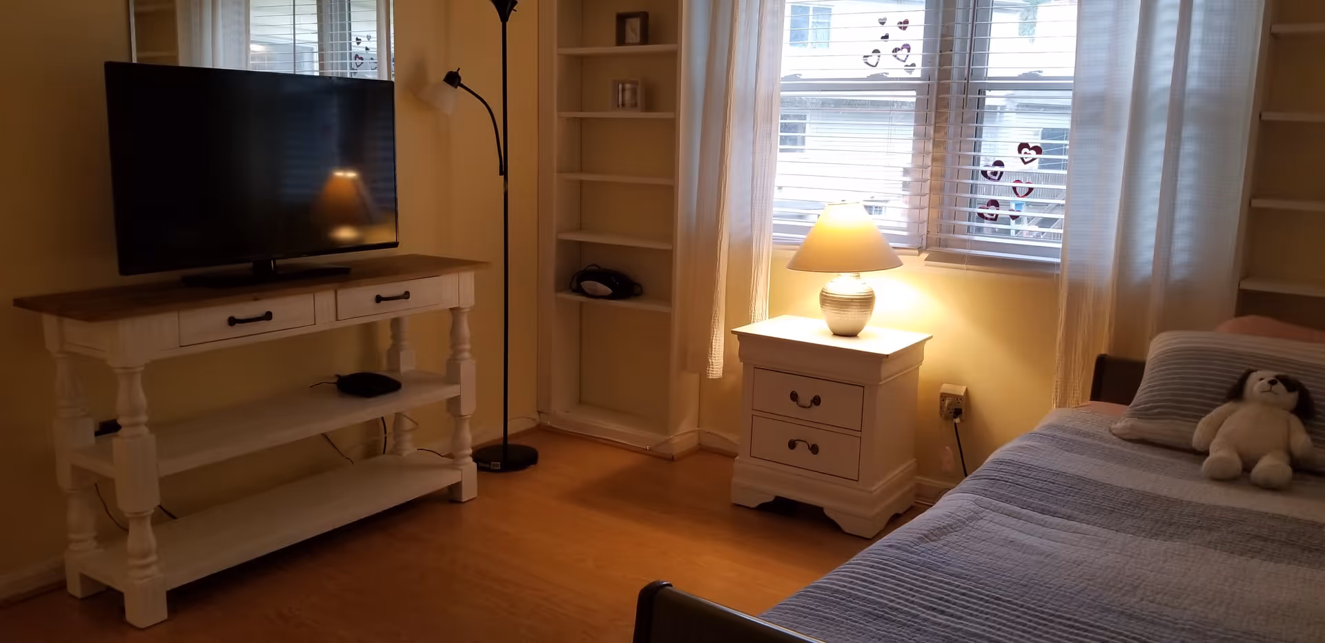 A cozy bedroom with a bed covered in a striped quilt and a stuffed dog toy on it. Next to the bed is a white nightstand with a lamp turned on. There is a window with white curtains and heart-shaped decorations on the glass. A white TV stand holds a flat-screen television, and a tall black floor lamp stands beside it. The room has wooden flooring and built-in shelves in the corner.