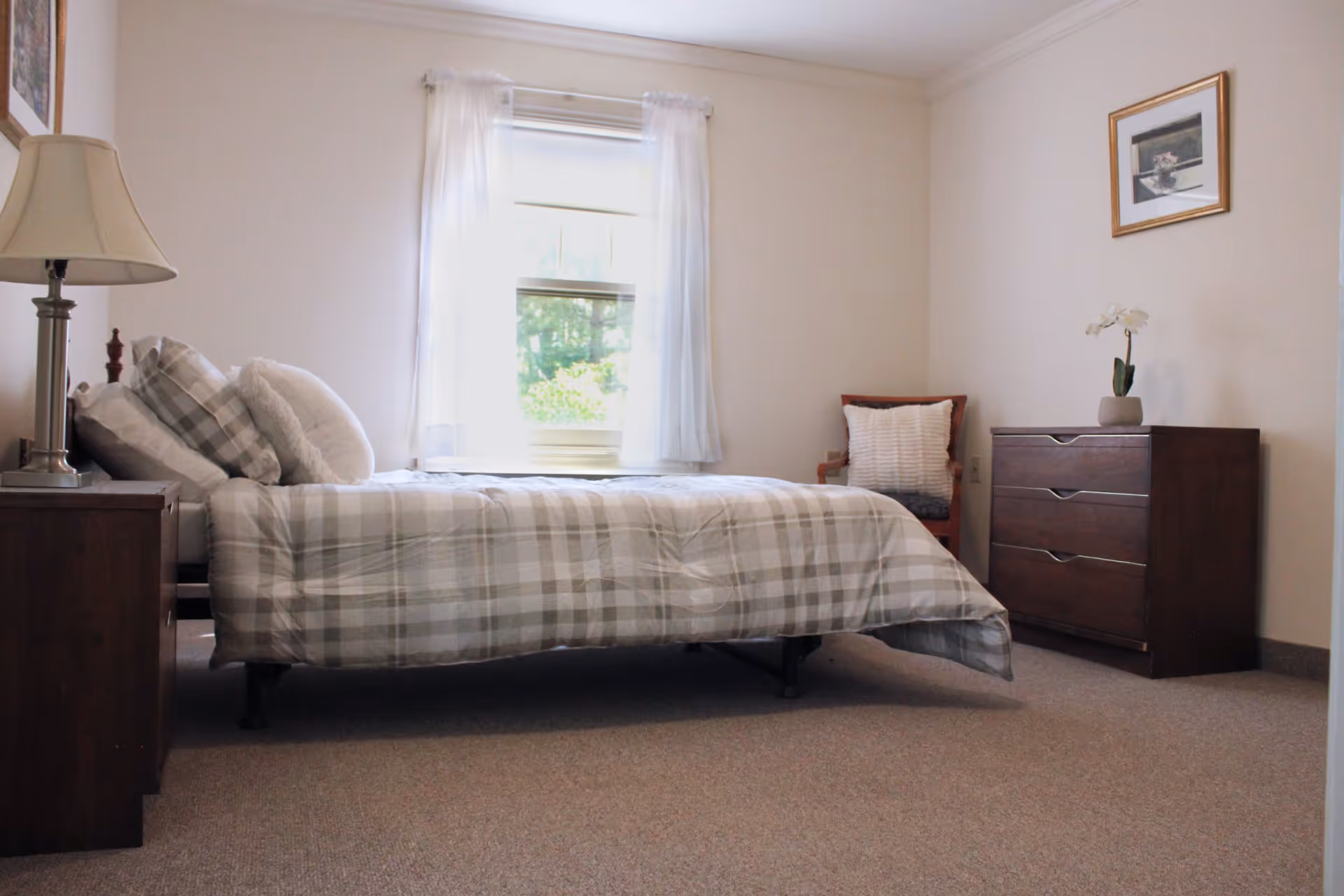 A simple, neatly arranged bedroom with a single bed covered in plaid bedding. The room has a window with sheer white curtains letting in natural light. There is a wooden nightstand with a lamp on the left side of the bed, a wooden dresser with a small plant on top on the right side, and a wooden chair with a cushion in the corner. A framed picture hangs on the wall above the dresser.