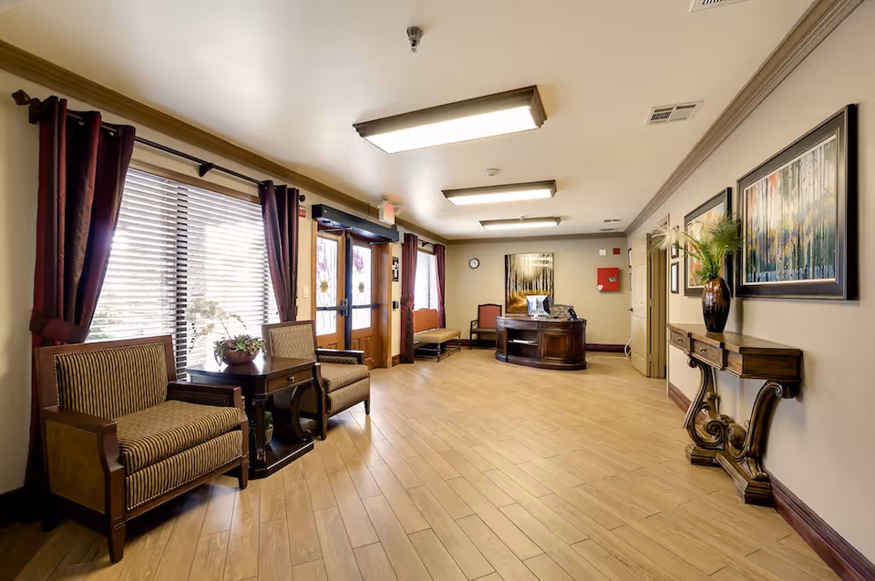 Well-lit senior living facility lobby with seating, a reception desk, artwork, and large windows with curtains.