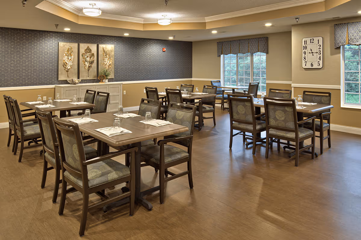 A dining room with several wooden tables and chairs arranged neatly. Each table is set with placemats, napkins, and upside-down drinking glasses. The room has large windows with patterned valances, a wall clock, and decorative artwork featuring leaves on one wall. The floor is wooden, and the lighting is warm and inviting.