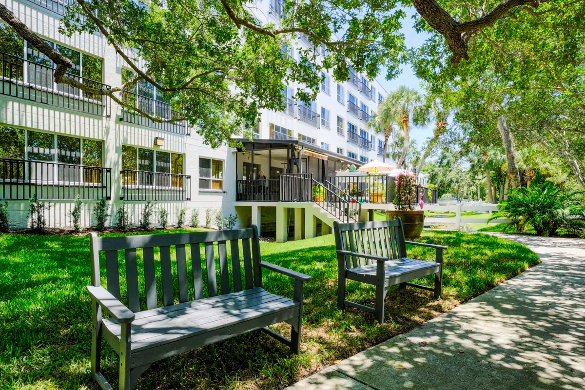 Wooden benches on a shaded grassy lawn beside a walkway in front of a multi-story senior living building with balconies and trees.