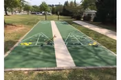 Two outdoor shuffleboard courts with painted scoring triangles separated by a central concrete walkway, surrounded by grass and trees.
