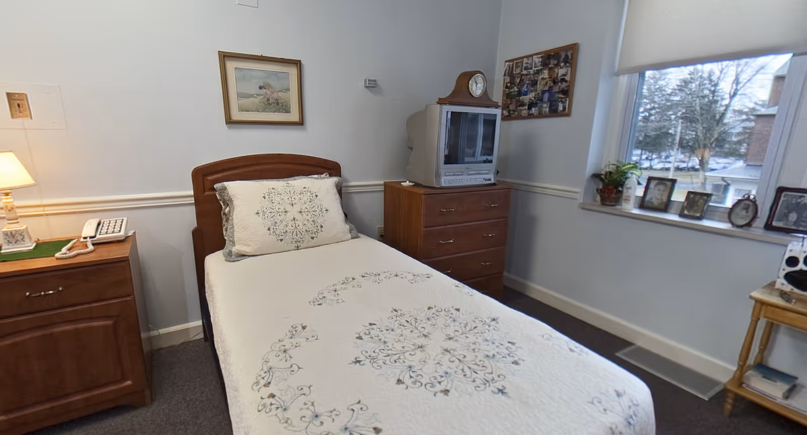 Small bedroom with a twin bed, wooden nightstand and dresser topped by a TV, and a window sill displaying framed photos.