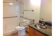 Bathroom with a white toilet, bathtub with shower and grab bars, and a sink with a granite countertop and wooden cabinet below. There is a large mirror above the sink and a towel rack on the wall.
