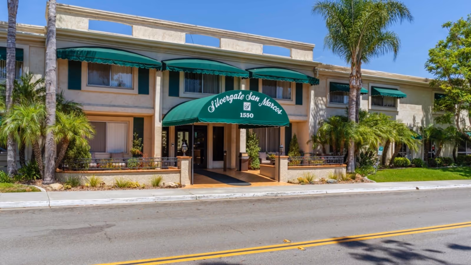 Front entrance of the Silvergate San Marcos building with green awnings, palm trees, and the street in front.