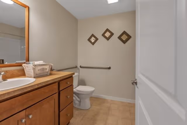 A bathroom with a wooden vanity cabinet topped with a sink and a large mirror above it. There is a basket with toiletries on the countertop. A white toilet is positioned against the far wall, which has three decorative square frames hanging above a metal grab bar. The floor is tiled, and the walls are painted beige. The bathroom door is partially open.