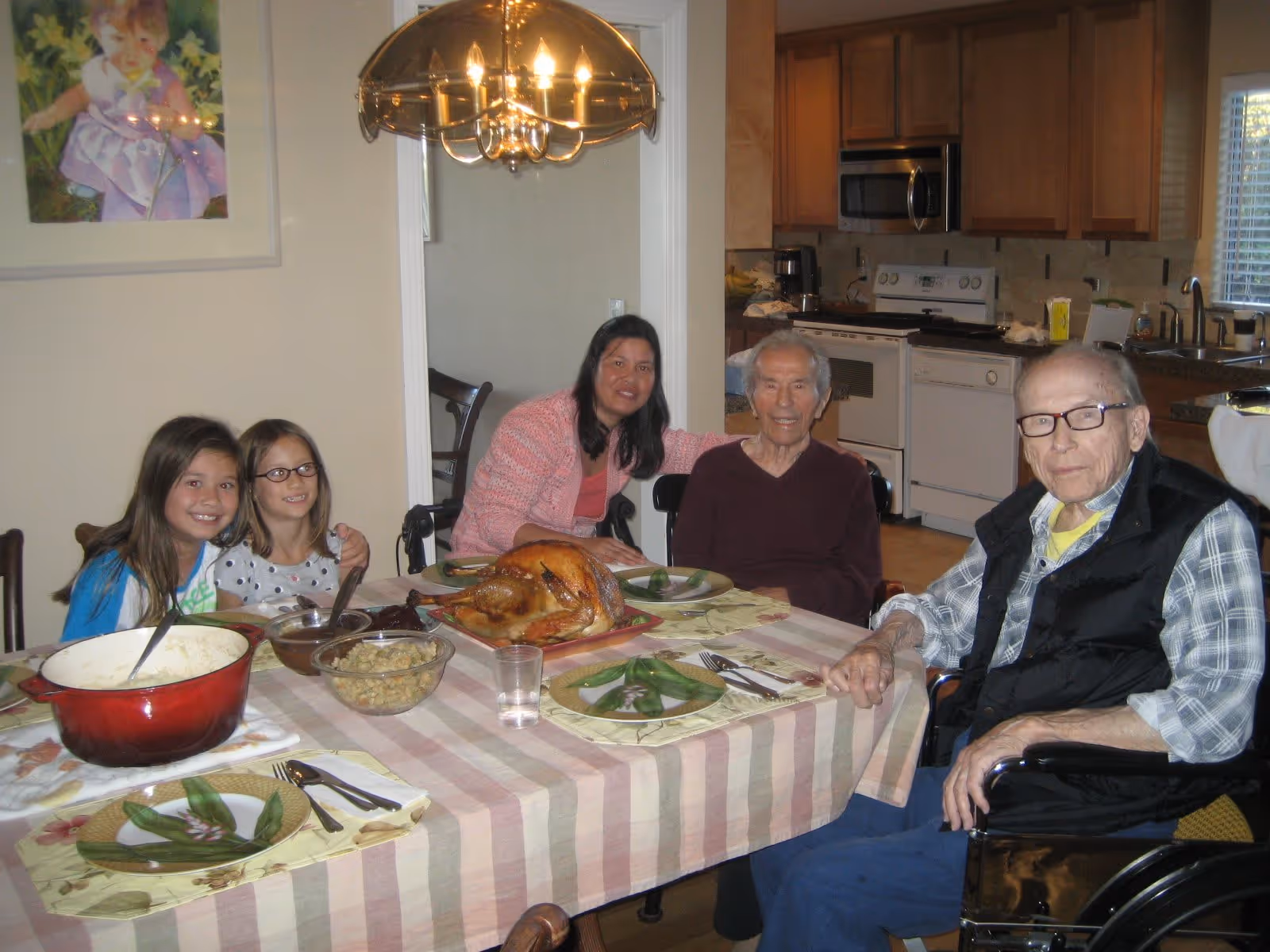 A family gathered around a dining table set with a roasted turkey, mashed potatoes, and other dishes in a home kitchen. Two elderly men and three younger individuals, including two children, are seated and smiling at the camera.