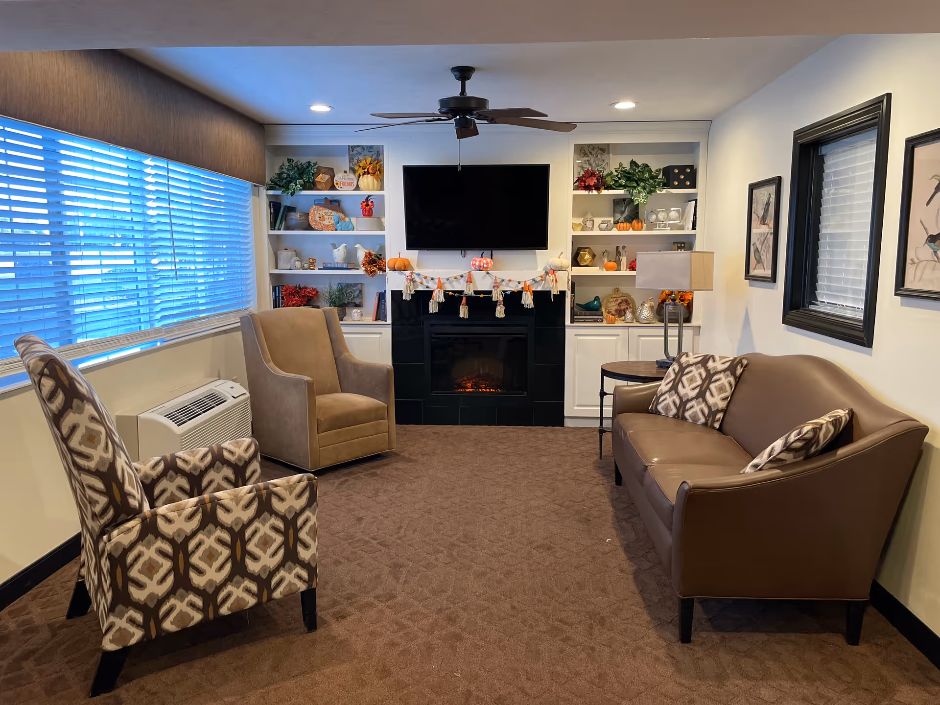 Cozy living room with patterned armchairs, a leather sofa, built-in shelves, a TV over a fireplace, and a large window with blinds.