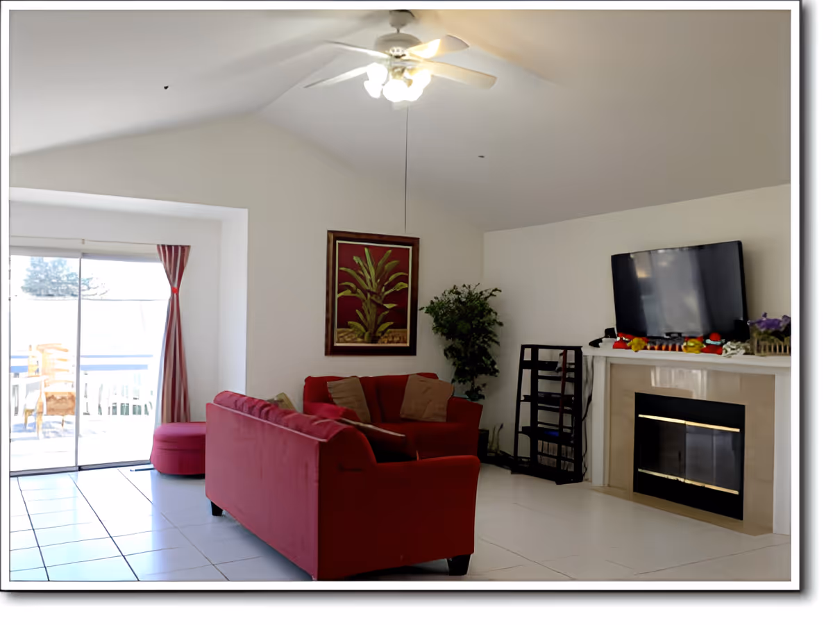 A bright living room with white walls and tiled floor featuring a red sofa set with cushions, a ceiling fan with lights, a framed plant artwork on the wall, a potted plant in the corner, a flat-screen TV mounted above a fireplace, and sliding glass doors with red curtains leading to an outdoor patio area.