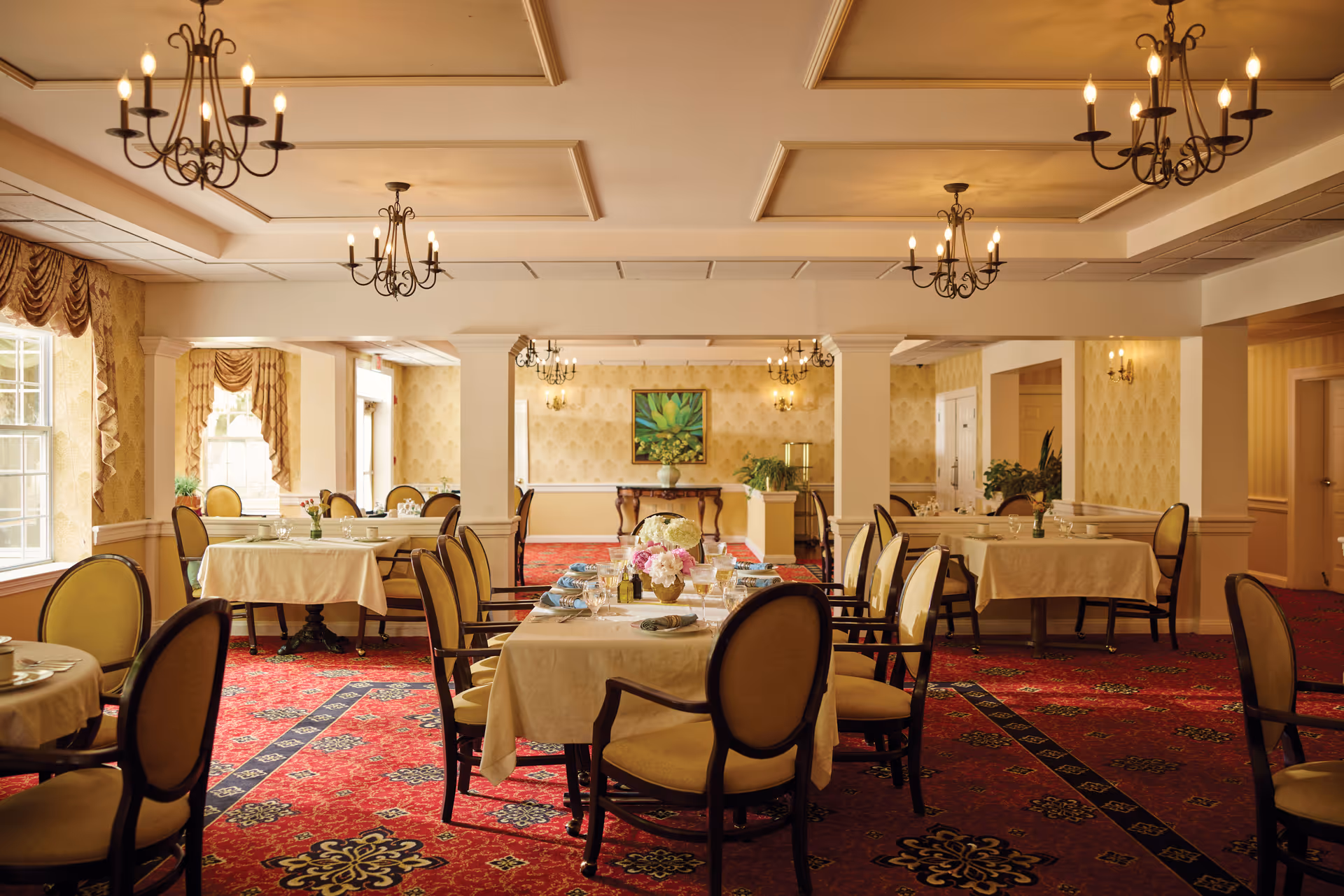 A warmly lit dining room with multiple tables covered in beige tablecloths, set with plates, glasses, and napkins. The room features upholstered chairs with wooden frames, red patterned carpet, chandeliers hanging from the ceiling, and large windows with draped curtains allowing natural light to enter. There is a decorative painting and plants on the far wall.