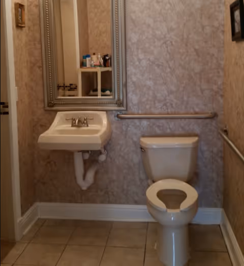 A small bathroom with a white toilet and a wall-mounted white sink. There is a large rectangular mirror with a decorative frame above the sink. The walls have a textured, marbled pattern, and there are metal grab bars installed next to the toilet and along the wall. The floor is tiled with beige tiles.