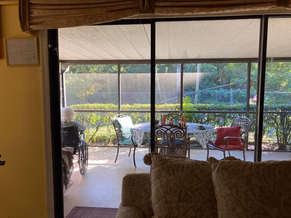 View from inside a living room looking out through large sliding glass doors onto a covered patio. On the patio, there is a round table with a patterned tablecloth and several chairs, one with a red cushion. An elderly person in a wheelchair is sitting on the left side of the patio, facing outside. Greenery and a chain-link fence are visible beyond the patio.