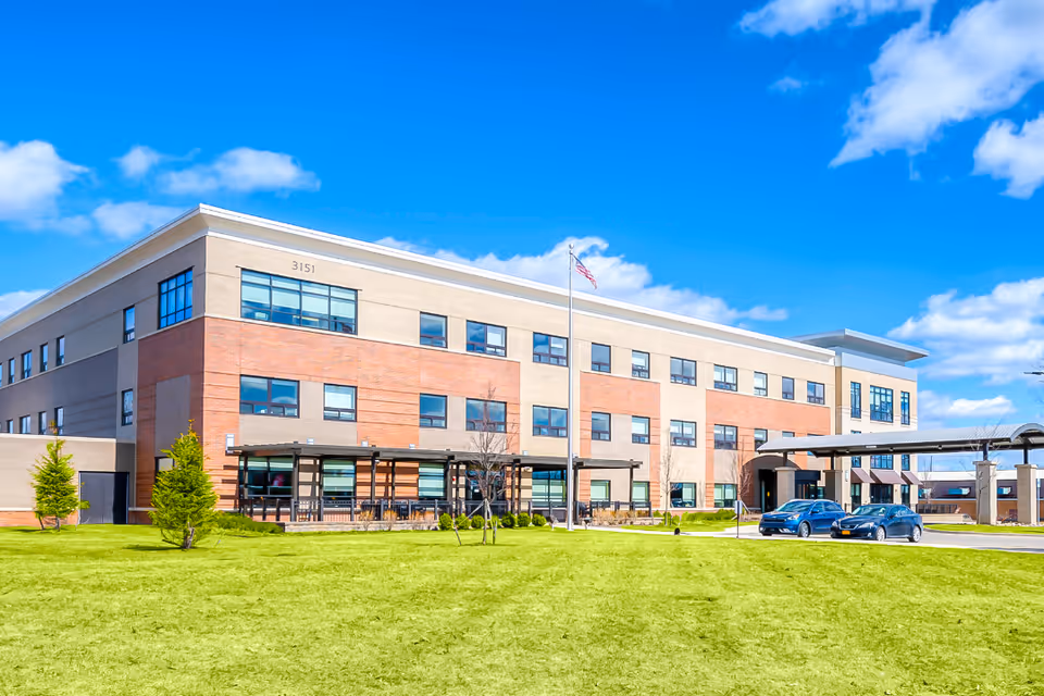 Exterior view of a modern three-story senior living facility building with a combination of beige and red brick facade. The building has many windows, a covered entrance with two cars parked nearby, a flagpole with an American flag, and a well-maintained green lawn in the foreground under a bright blue sky with scattered clouds.