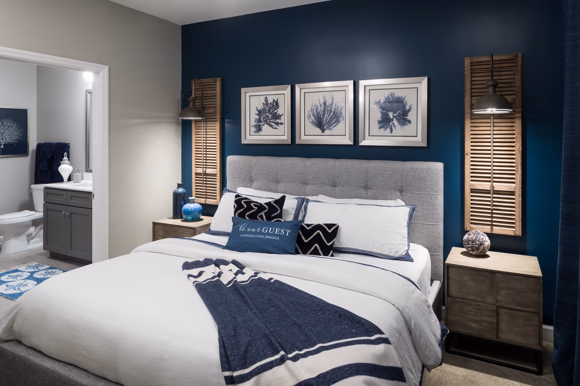 A modern bedroom with a large gray upholstered bed featuring white and navy blue bedding and decorative pillows, including one that says 'be our GUEST AVENIDA COOL SPRINGS'. The wall behind the bed is painted dark blue and decorated with three framed botanical prints and two wooden shutter-style wall sconces with lamps. A wooden nightstand with decorative items is on each side of the bed. An open door reveals a bathroom with a toilet, vanity, and towel rack.