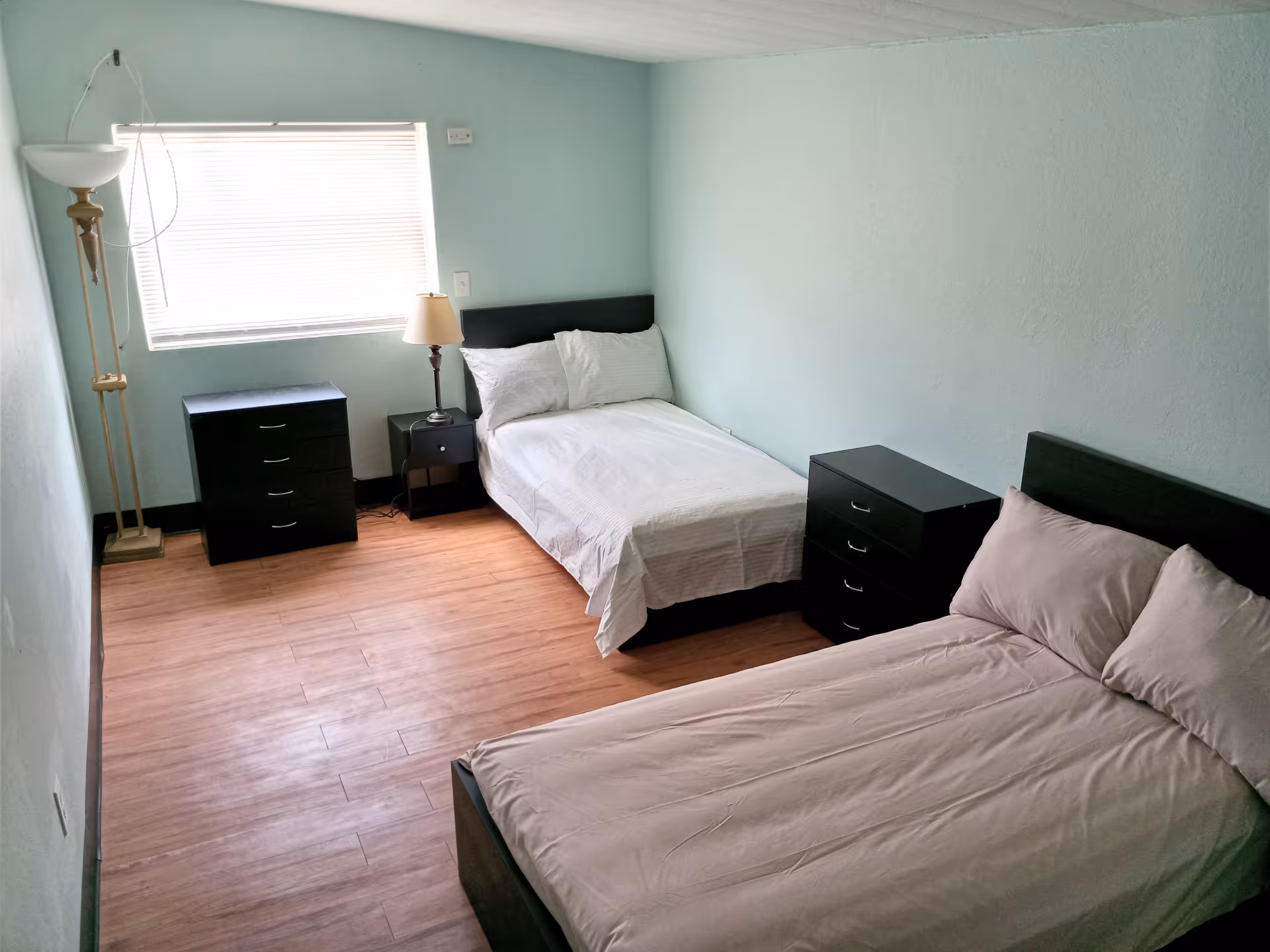 A simple bedroom with two beds, one with white bedding and the other with light pink bedding. The room has light blue walls, a wooden floor, a window with blinds, a floor lamp, a table lamp on a small nightstand, and two black dressers.