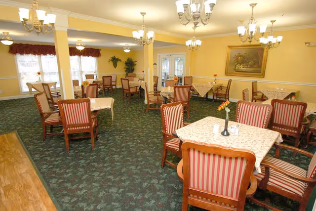 A spacious dining room with multiple tables covered in patterned tablecloths, each surrounded by wooden chairs with red and beige striped upholstery. The room has yellow walls, green patterned carpet, chandeliers hanging from the ceiling, and large windows with red valances allowing natural light to enter. A framed painting and some plants decorate the walls.