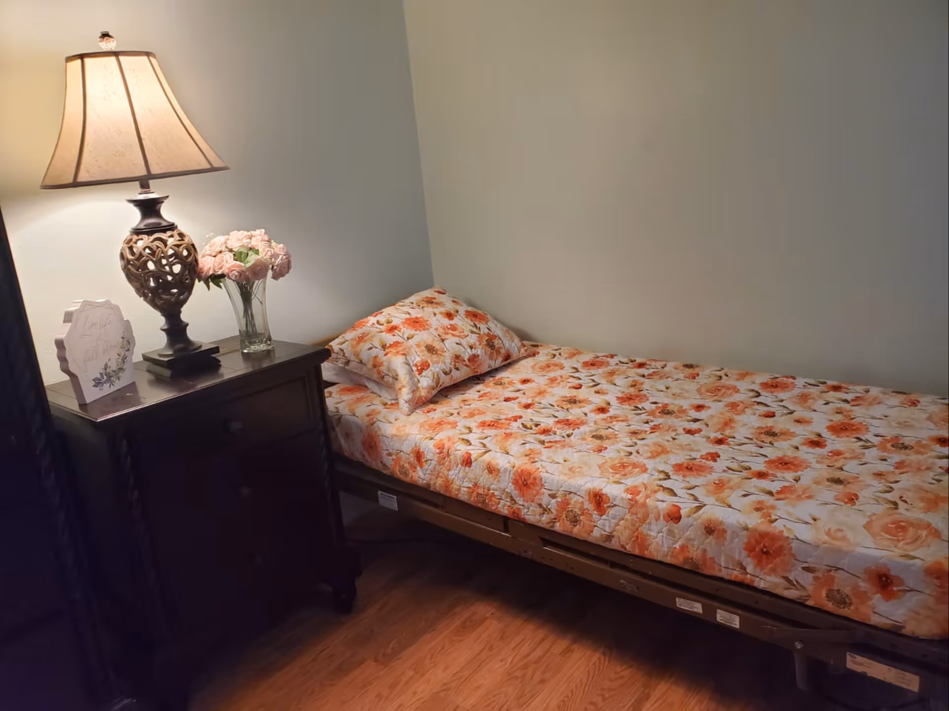 A small bedroom with a single bed covered in a floral-patterned bedspread and matching pillow. Next to the bed is a dark wooden nightstand with three drawers, a decorative lamp, a vase of pink flowers, and a small framed sign. The room has light-colored walls and wooden flooring.