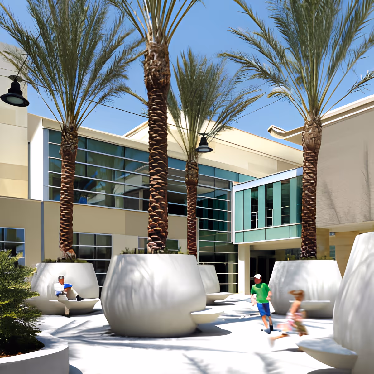 Outdoor courtyard area with large white planters containing tall palm trees. Modern building with large windows surrounds the courtyard. Two children are running and a person is sitting on one of the planter benches.