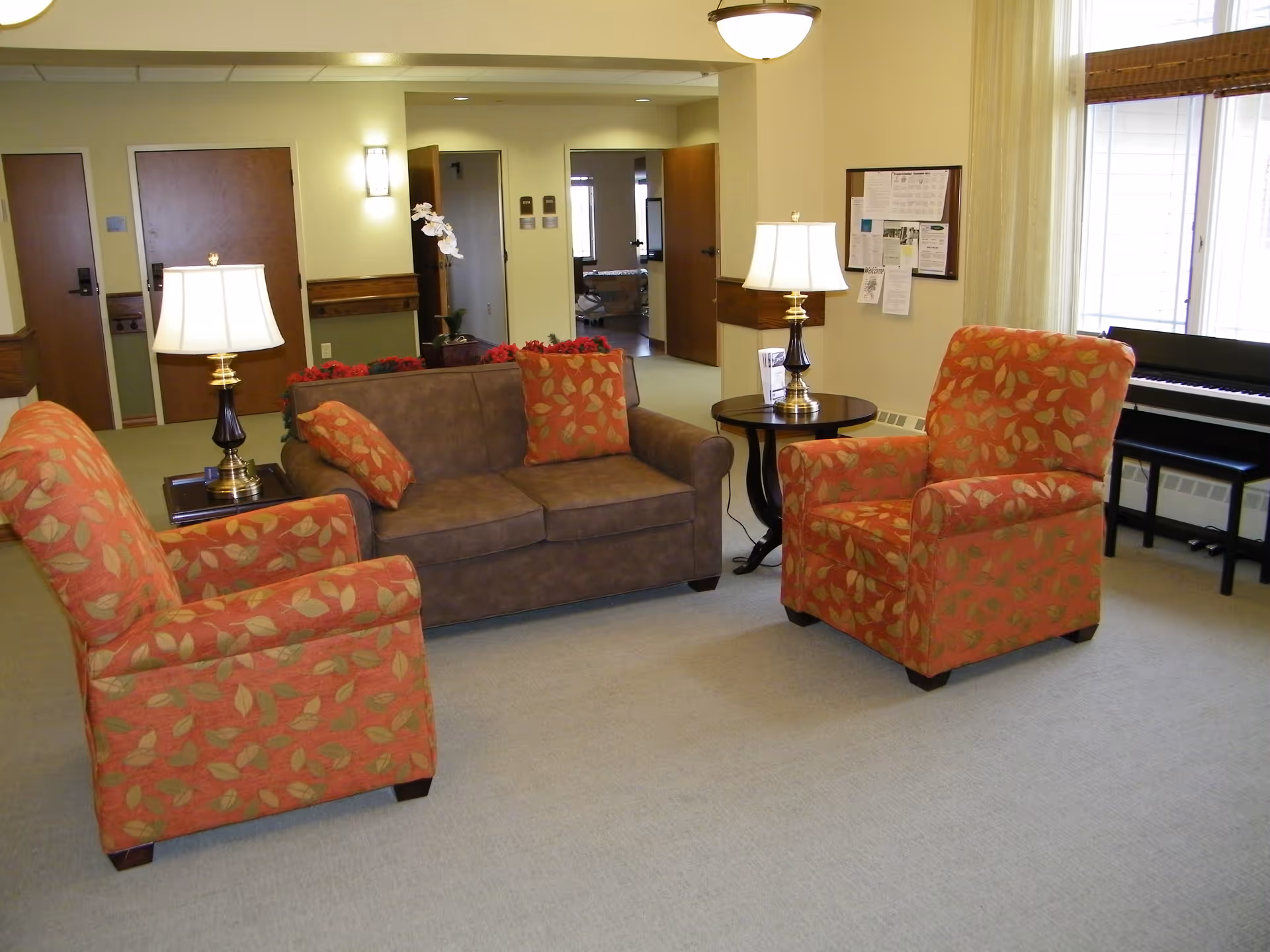 A cozy living room area in a senior living facility with a brown sofa and two orange armchairs with leaf patterns. There are two table lamps on side tables, a bulletin board on the wall, and a piano near a window with sheer curtains. Several doors and a hallway are visible in the background.