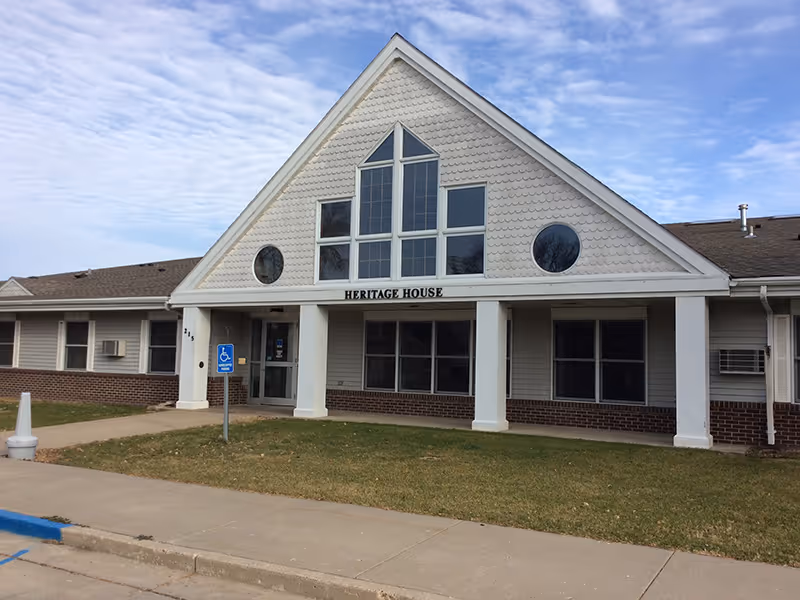 Exterior front view of a single-story building named Heritage House with a triangular roof section featuring multiple windows, a brick lower facade, white columns at the entrance, and a handicap parking sign on the lawn.
