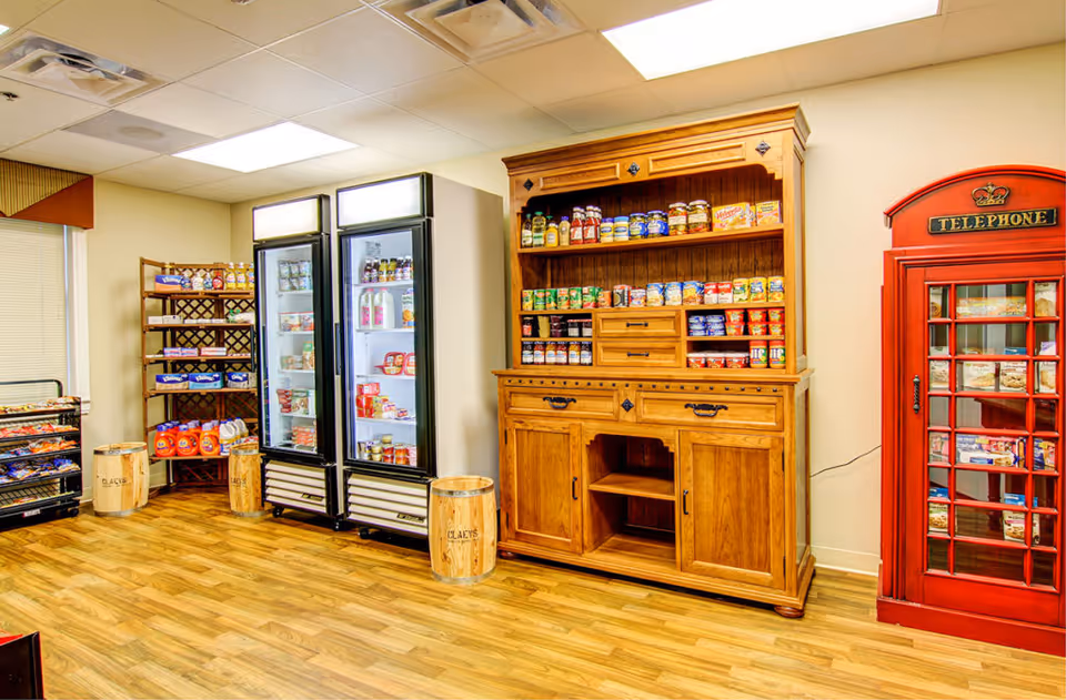 Interior view of a small convenience area with wooden flooring, featuring two glass-door refrigerators stocked with various food items, a wooden cabinet filled with canned goods and condiments, shelves with snacks and other packaged foods, and a red British-style telephone booth repurposed as a display case.