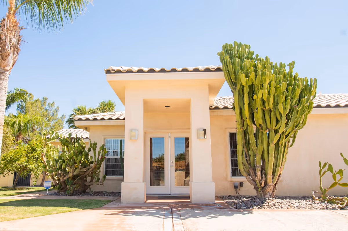 Front exterior view of a single-story building with a tiled roof, beige stucco walls, and a central entrance with double glass doors. There are large cacti and other desert plants on either side of the entrance, and a clear blue sky overhead.