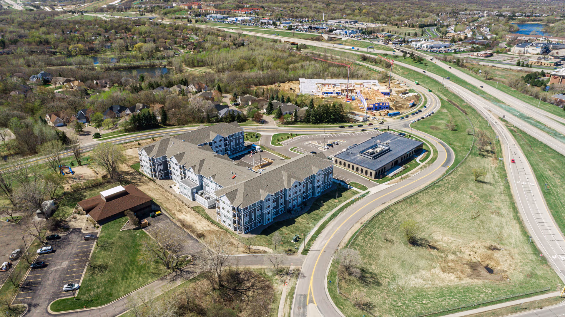 Aerial view of Havenwood of Burnsville senior living facility showing a large multi-wing building with a parking lot, surrounded by roads, grassy areas, and nearby residential neighborhoods. There is also a smaller building adjacent to the main facility and a highway visible in the background.
