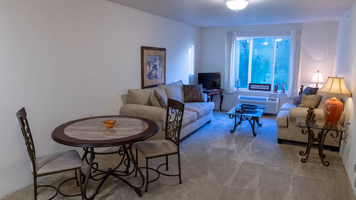 A cozy living room with two beige sofas, a glass coffee table, and a small round dining table with two chairs. The room has a window with white curtains, a TV on a wooden stand, and two side tables with lamps and decorative figurines. The walls are plain white and the floor is carpeted.