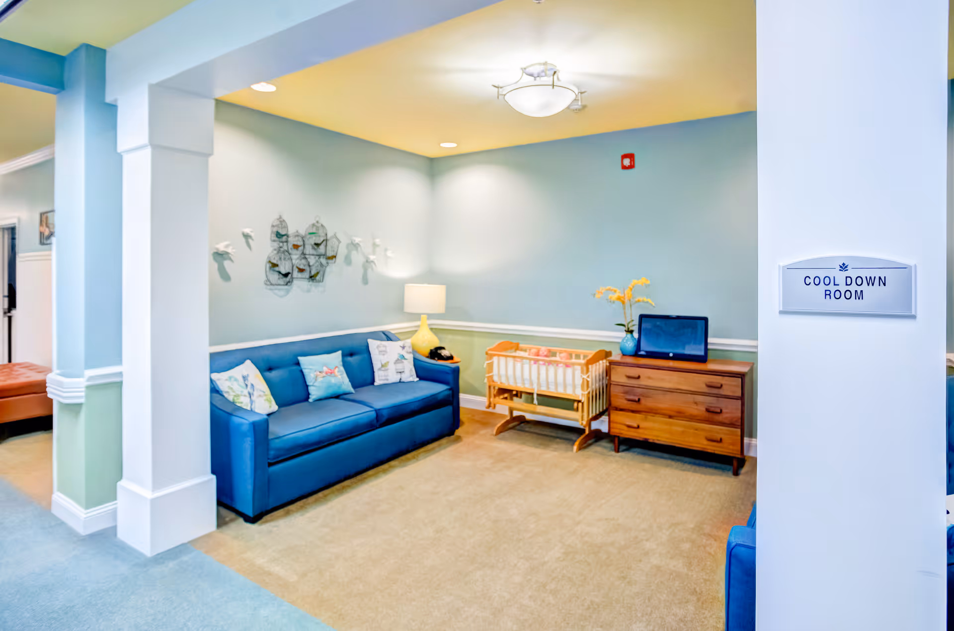 A cozy room labeled as 'Cool Down Room' with a blue couch adorned with decorative pillows, a wooden dresser with a vase of yellow flowers and a laptop on top, a small wooden cradle, and a wall decoration featuring birds and birdcages. The walls are painted light blue with white trim, and the room is softly lit by a ceiling light and a table lamp.