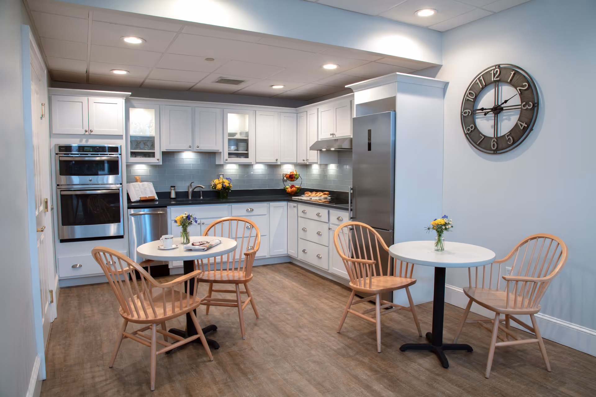 A modern kitchen area with white cabinetry, stainless steel appliances including a refrigerator and double oven. Two small round tables each surrounded by three wooden chairs are placed on a wooden floor. Each table has a small vase with flowers. A large wall clock is mounted on the light blue wall.