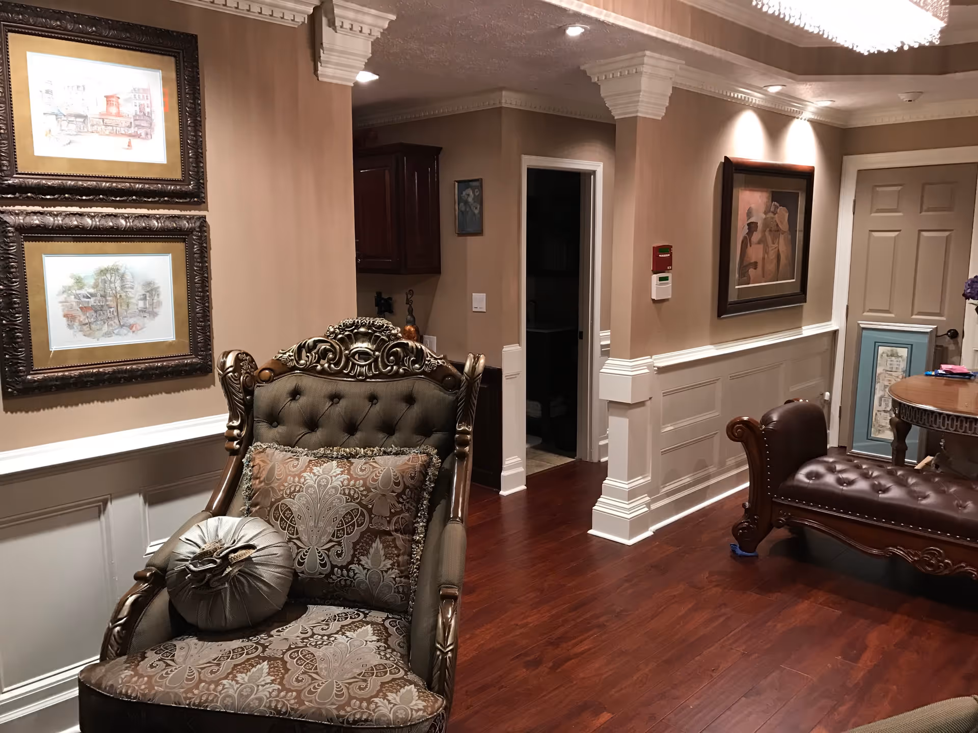 A cozy interior room with classic decor featuring a tufted armchair with patterned cushions, a dark wooden chaise lounge, framed artwork on beige walls, and polished hardwood floors. The room has white wainscoting and crown molding, with a doorway leading to another room.