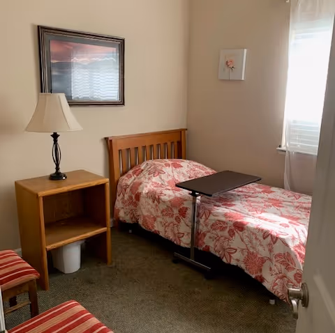 Small bedroom with a single bed covered in a red floral quilt, a wooden bedside table with a lamp, an overbed table, two striped chairs, and a window with sheer curtains.