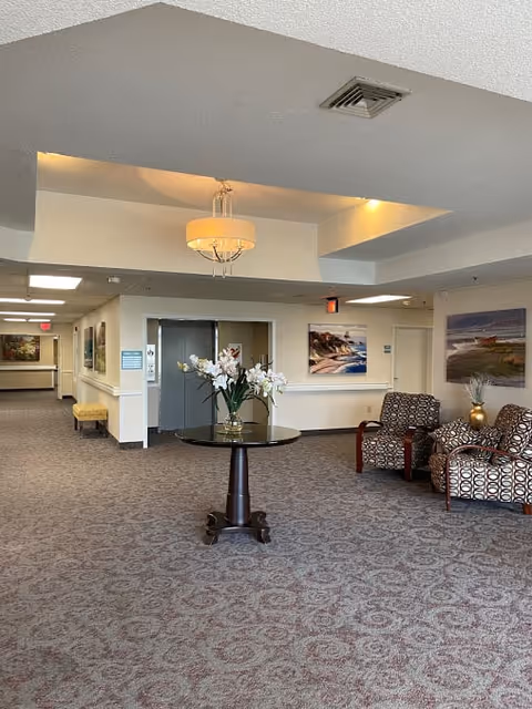 Interior view of a senior living facility hallway with patterned carpet, a round table with a vase of white flowers in the center, two patterned armchairs with a small table and decorative vase, and framed landscape paintings on the walls. The ceiling has recessed lighting and a hanging light fixture.