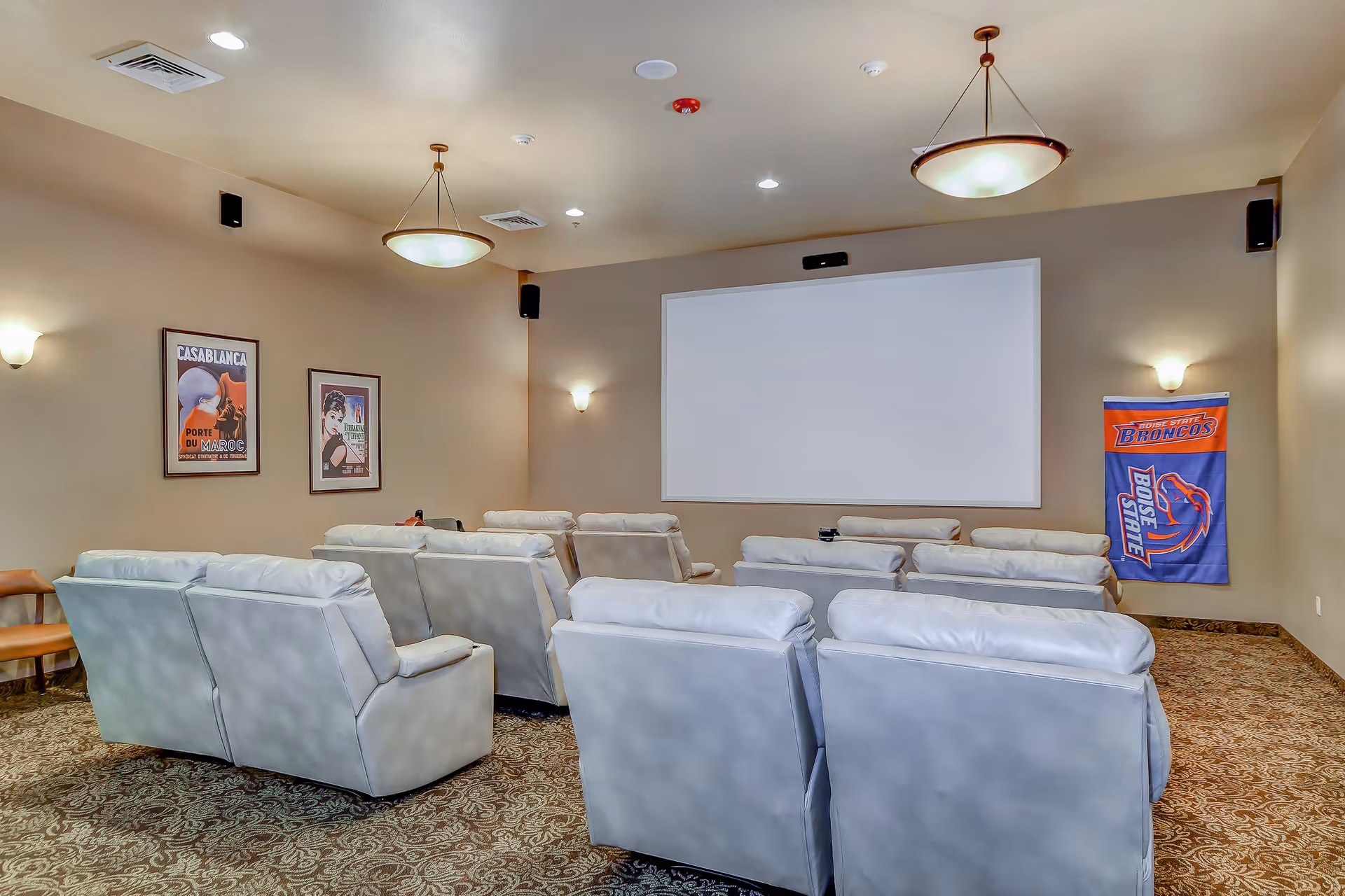 A cozy movie theater room with multiple rows of white cushioned recliner chairs facing a large blank projection screen. The walls are beige with framed vintage movie posters and a Boise State Broncos banner. The room is softly lit with ceiling and wall lights.