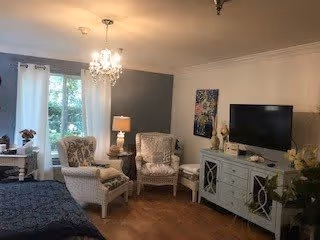 A cozy living room with two patterned armchairs, a small round table with a lamp between them, a chandelier hanging from the ceiling, a large window with curtains, a flat-screen TV on a light-colored cabinet, and decorative artwork on the walls.