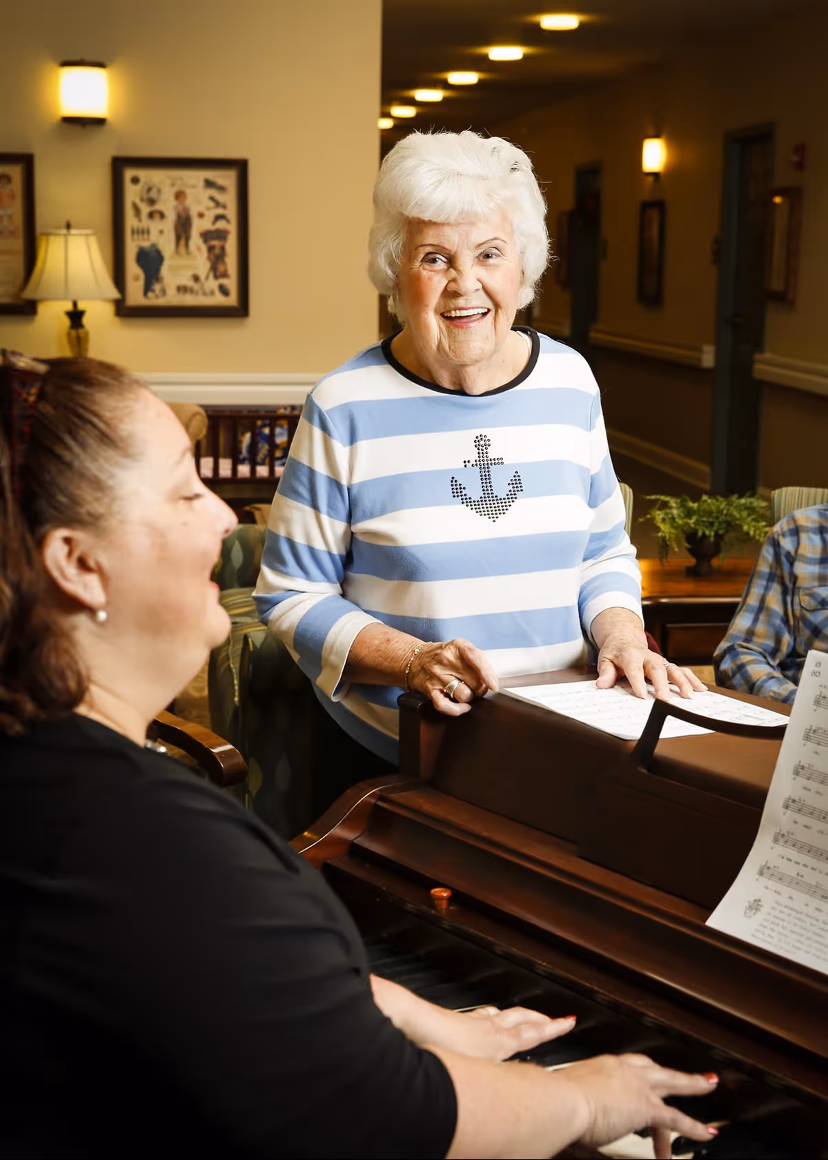 An elderly woman wearing a blue and white striped shirt with an anchor design stands smiling near a piano while another woman plays the piano in a warmly lit room with framed pictures and a lamp in the background.