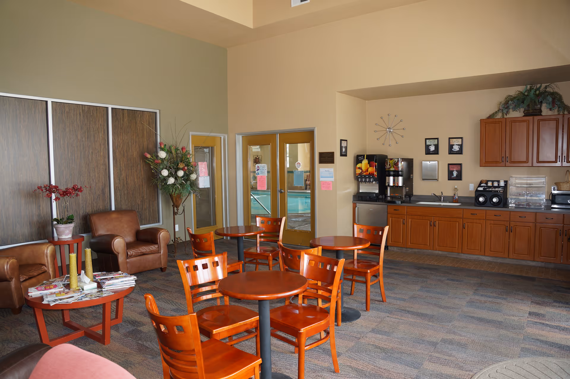 A small communal lounge with round wooden tables and chairs, a kitchenette/coffee station on the right, and leather armchairs and plants on the left.